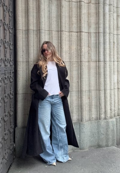 Mujer con cabello largo y rubio que lleva gafas de sol, camisa blanca, abrigo largo negro y vaqueros anchos azules apoyada contra una pared de columnas de piedra.