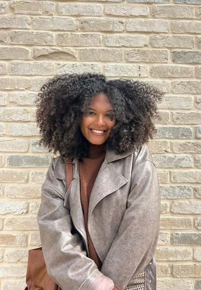 Chaqueta de piel sintética gris con cuello ancho, sobre un suéter de punto marrón. Rizos texturizados enmarcan la cara, bolso marrón colgado del hombro.