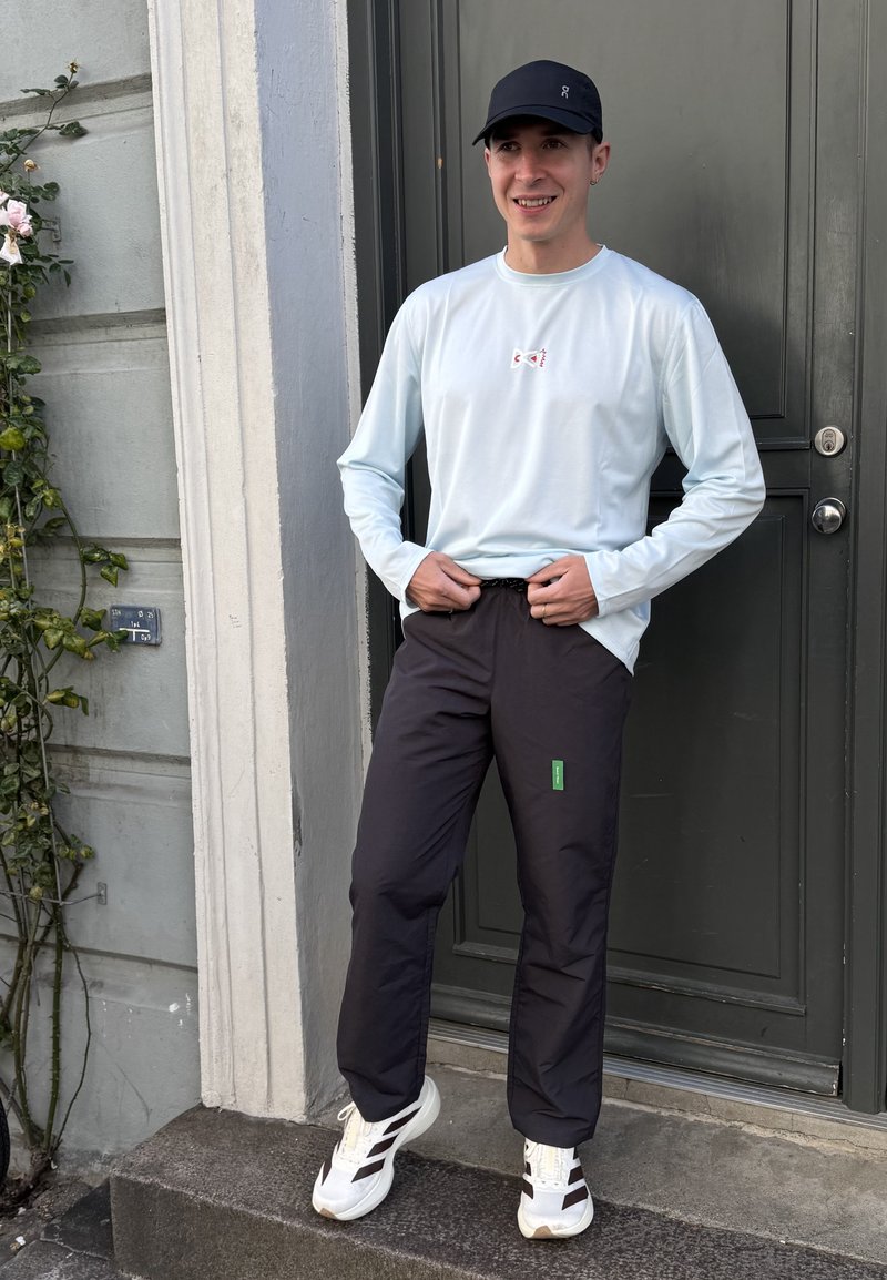 Joven con gorra negra, camisa de manga larga azul claro, pantalones negros y zapatillas blancas de pie junto a una puerta gris oscuro con flores en la pared.