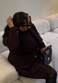 Blazer supradimensionat maro-roșcat cu nasturi aurii, purtat peste un top negru, asortat cu o geantă neagră matlasată și colanți negri cu model.