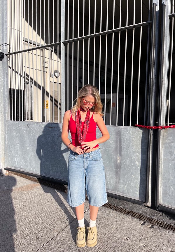 Joven mujer con camiseta sin mangas roja y bufanda, pantalones cortos de mezclilla, zapatos beige, de pie al aire libre frente a una gran puerta metálica bajo la luz del sol.