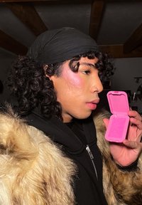 Lockigt svart hår, iklädd svart huvudduk och en pälsjacka. Håller en rosa sminkväska, strukturerad med ett geometriskt mönster.