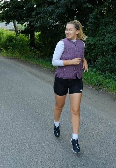 Corredor vestido con un chaleco acolchado morado y pantalones cortos negros, con calcetas blancas y zapatos negros, en un camino de grava rodeado de vegetación.