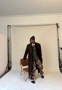 Muž v černé čepici, slunečních brýlích, dlouhém tmavém kabátě, zeleném saku, červené kravatě a maskáčových kalhotách stojí vedle dřevěné židle ve fotografickém studiu.