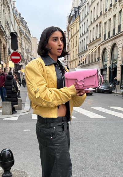 Mujer con chaqueta amarilla y pantalones negros que sostiene un bolso rosa, parada en un paso de peatones en una zona urbana con edificios y peatones.