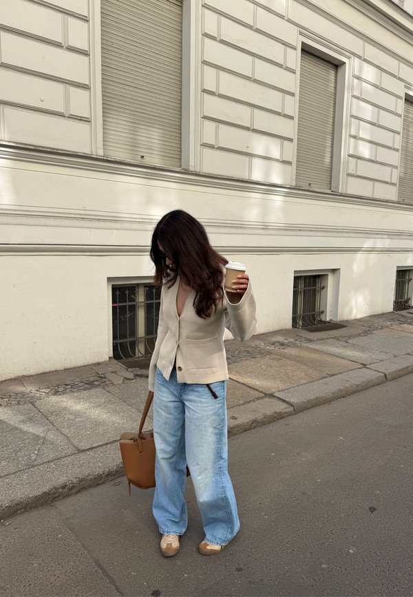 Woman with long dark hair wearing beige cardigan and wide blue jeans, holding a coffee cup and brown handbag on a city sidewalk.