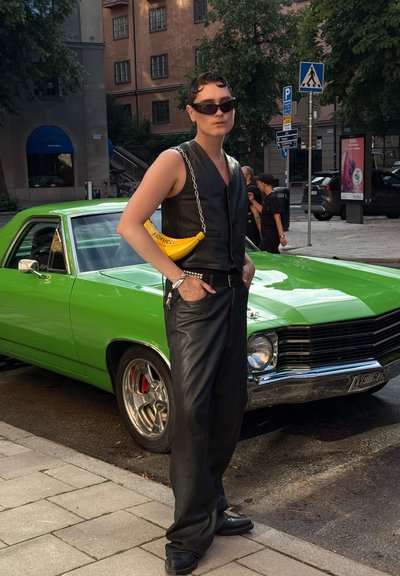 Persona con chaleco y pantalones de cuero negro, gafas de sol y un bolso amarillo en forma de plátano se encuentra al lado de un coche clásico de color verde brillante en una calle de la ciudad.