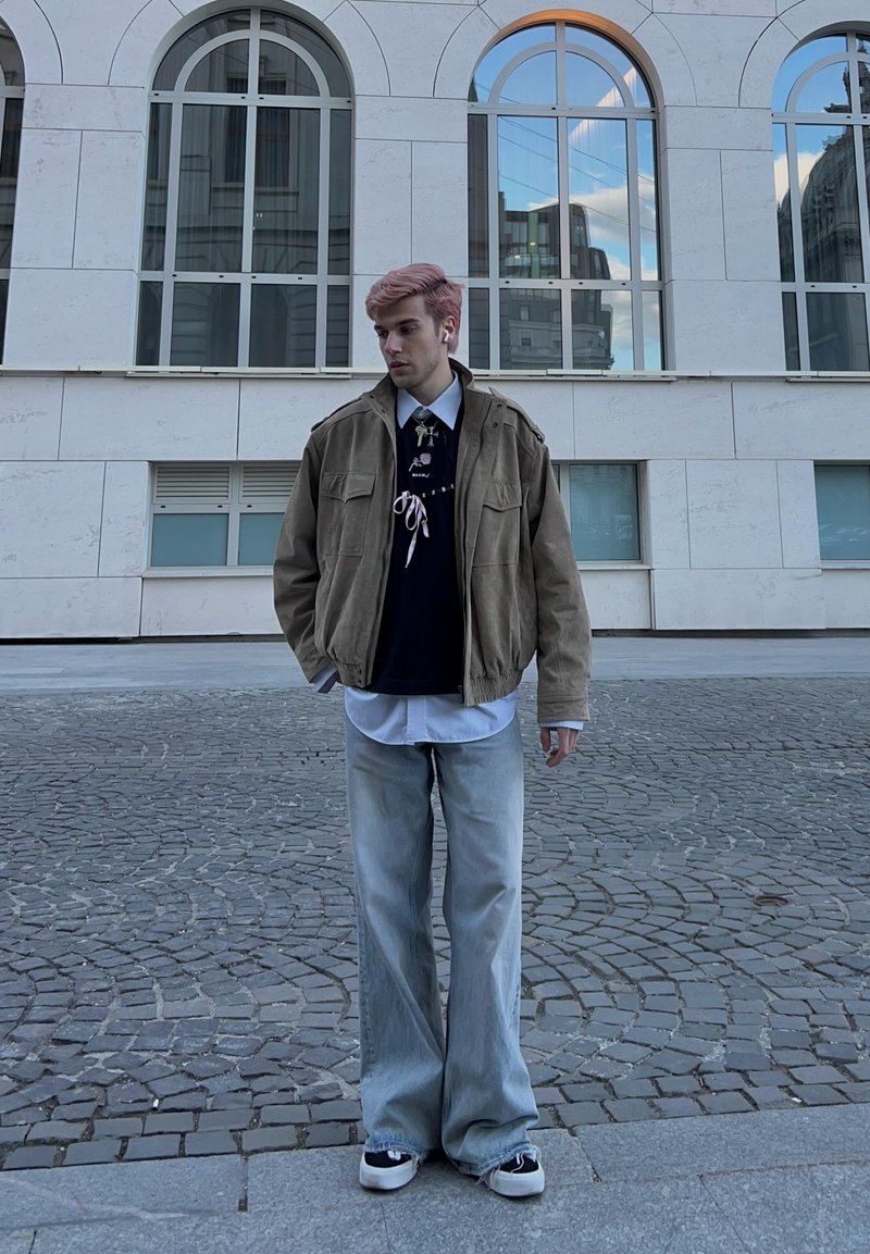 Jeune homme aux cheveux roses portant une veste marron, des chemises superposées, un jean bleu clair et des baskets, debout sur un pavé près d'un bâtiment.