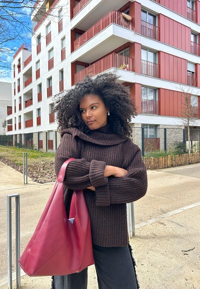 Suéter marrón de canalé con un cuello alto, combinado con un gran bolso de cuero burdeos. Entorno urbano con edificios de apartamentos modernos al fondo.