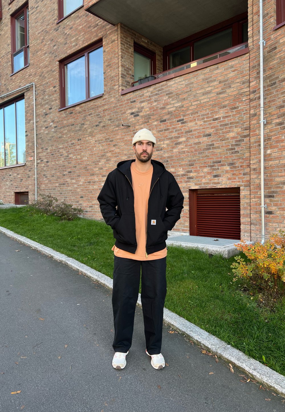 Black zip-up hoodie over an orange t-shirt, black trousers, and white trainers. A beige beanie completes the outfit against a brick wall.