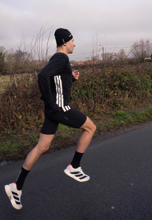 Corridore maschio in abbigliamento sportivo nero e cappello che corre all'aperto su una strada asfaltata accanto a cespugli senza foglie sotto un cielo nuvoloso.
