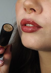 Leppepenn med svart ribbet design og gullaksenter, holdt nær lepper malt i en blank rødbrun nyanse, med en gull septumring.