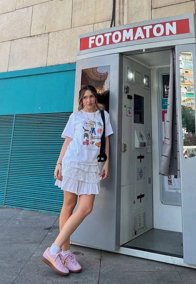 Camiseta blanca con gráfico colorido, falda blanca con capas, zapatillas rosas y bolso negro; posicionada junto a una cabina de fotos con detalles rojos y plateados.