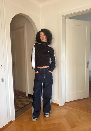 Joven con cabello rizado parada en una puerta, lleva una chaqueta negra recortada, jeans oscuros de pierna ancha con franjas laterales y zapatillas grises.