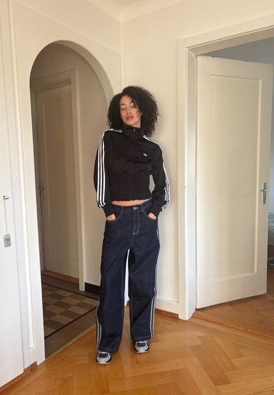 Joven con cabello rizado parada en una puerta, lleva una chaqueta negra recortada, jeans oscuros de pierna ancha con franjas laterales y zapatillas grises.