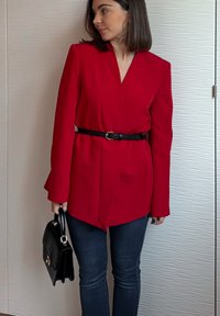 Blazer rosso con design su misura, maniche lunghe e scollo a V, stretto in vita con una cintura nera. Abbinato a jeans scuri e una borsa nera.