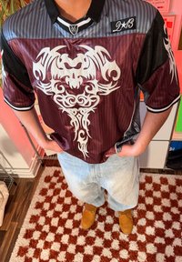 Personne portant un t-shirt bordeaux et gris avec un motif de crâne tribal blanc, un jean bleu clair et des chaussures beige, debout sur un tapis à carreaux.
