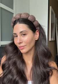 Bandeau en tissu marron avec des accents arrondis. Texture douce, finition lisse. Cheveux lâchement coiffés, maquillage naturel avec des imperfections minimales visibles.