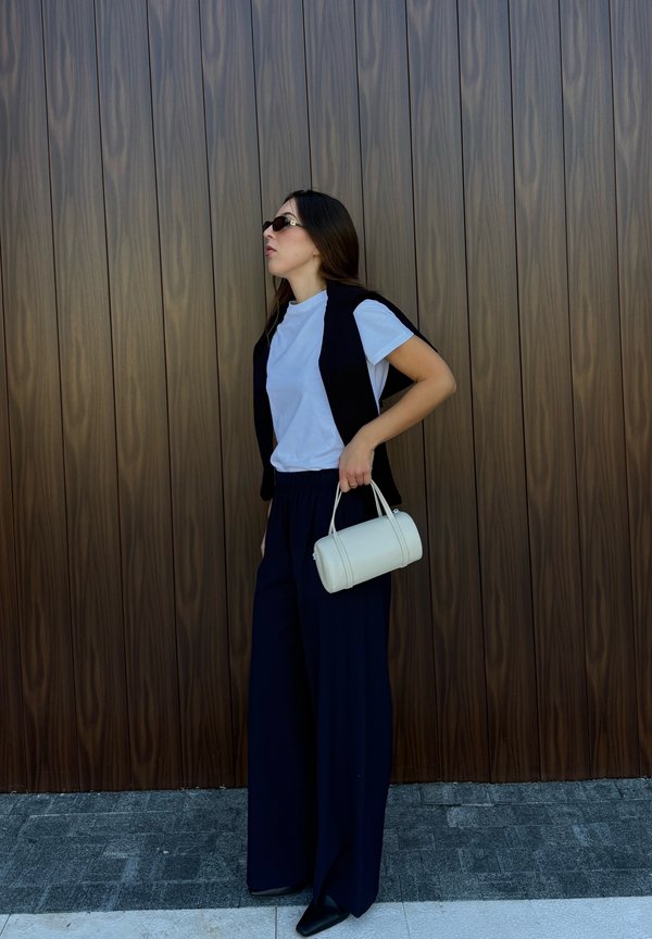Una mujer con gafas de sol, una camiseta blanca, un chaleco negro y unos pantalones de pernera ancha sostiene un bolso blanco junto a una pared de madera.