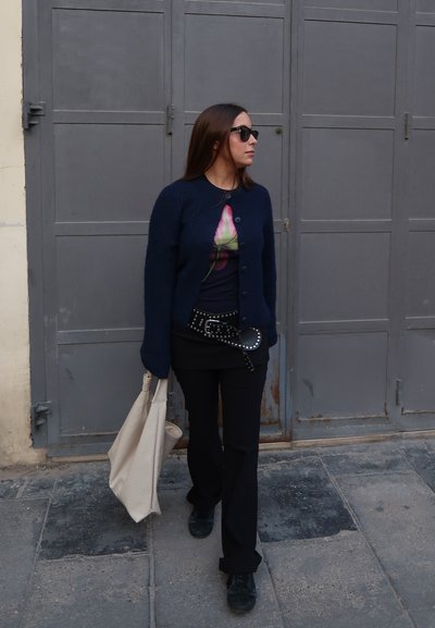 Cárdigan de punto azul marino oscuro, camiseta gráfica con flores, pantalones negros, cinturón con tachuelas y bolso de lona gris. Las gafas de sol completan el atuendo.