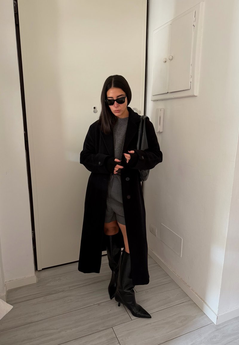 Casaco comprido preto sobre um suéter cinza e calções, combinado com botas pretas até ao joelho. Os acessórios incluem óculos de sol pretos e uma mala preta.