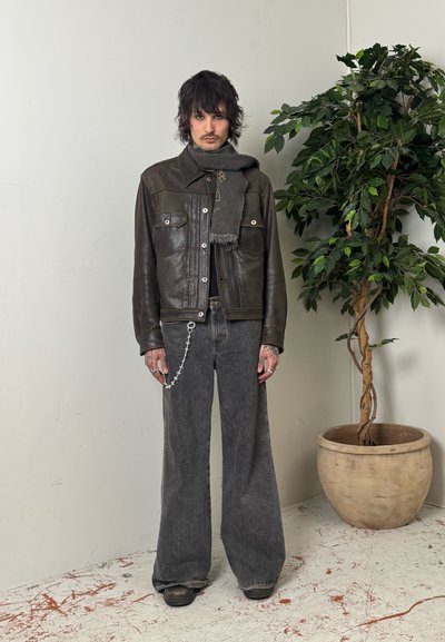 Joven con cabello oscuro y despeinado, vistiendo una chaqueta de cuero oscura, bufanda gris, jeans anchos grises con cadena, de pie junto a una planta frondosa en maceta.