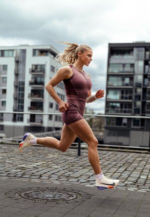 Joven mujer con ropa deportiva corriendo por una calle empedrada en una zona urbana con modernos edificios de apartamentos al fondo.