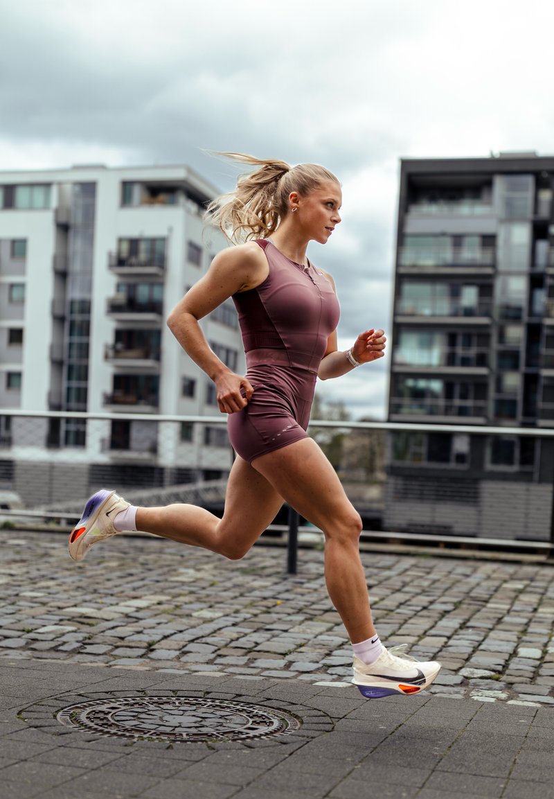 Jeune femme en tenue de sport courant sur une rue pavée dans une zone urbaine avec des bâtiments modernes en arrière-plan.