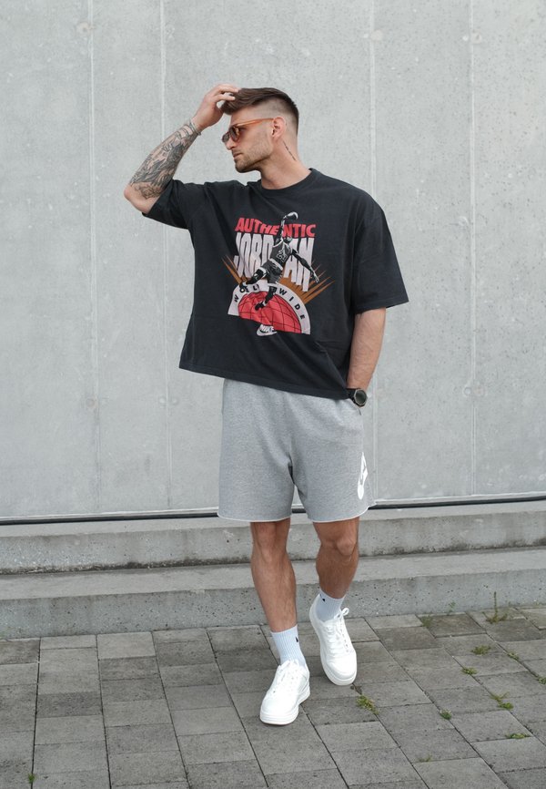 Muž v slunečních brýlích, který nosí černé tričko s grafikou Jordan, šedé šortky značky Nike, bílé tenisky a bílé ponožky, stojí u betonové zdi.