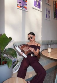 Osoba sedící na světle modrém polštáři, oblečená v tmavě fialovém off-shoulder outfitu, čte časopis, vedle ní je zelená rostlina a nápoj.