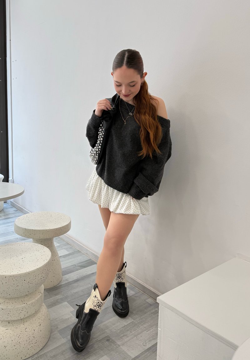 Jeune femme portant un pull épaules dénudées foncé, une jupe blanche à pois, des bottes noires et blanches, tenant un sac clouté, debout à l'intérieur près d'un mur blanc.