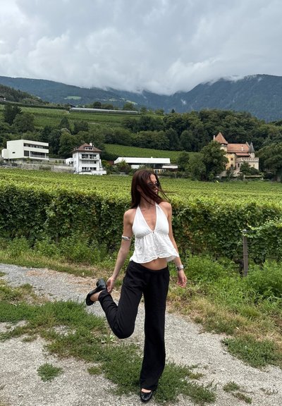 Top blanco de tirantes con dobladillo en volantes, pantalones negros holgados y zapatos negros. Entorno al aire libre con viñedos verdes y montañas nubladas al fondo.
