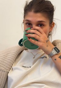 Žalias keraminis puodelis laikomas rankose, vilkint baltą marškinėlį su juodu tekstu, mezginį smėlio spalvos megztinį ir sidabrinį skaitmeninį laikrodį.