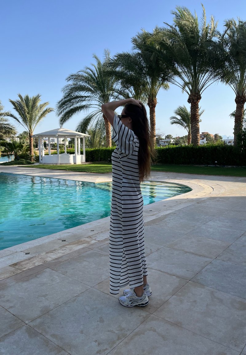 Ρυθμισμένο μακρύ φόρεμα σε λευκό και μαύρο, με κοντά μανίκια και φαρδιά εφαρμογή. Συνδυάζεται με γκρι αθλητικά παπούτσια. Πίσω βάθος πισίνα και φοίνικες.