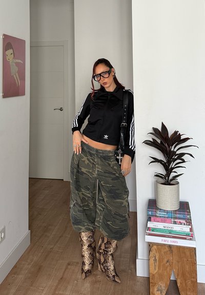 Mujer con chaqueta negra de Adidas, pantalones cargo de camuflaje y botas de piel de serpiente, de pie en el interior junto a una planta sobre libros apilados.