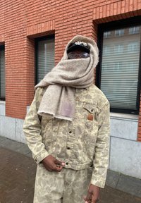 Μπεζ τζιν μπουφάν με σχέδιο και ταιριαστά παντελόνια, τυλιγμένο σε μια μεγάλη ανοιχτό καφέ κασκόλ, με μια ετικέτα ορατή στην τσέπη του μπουφάν.