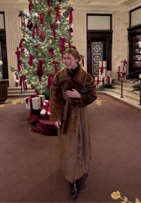 Brun faux fur-frakke med bælte i taljen, oversize ærmer og knapper. Baggrunden viser et juletræ med pynt og gaver.