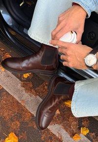 Person som sitter i bil med brune lærstøvler og lyseblå jeans, føttene ute av bilen, holder en hvit takeaway kaffekopp. Høstblader på bakken.