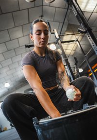 Idrettsutøver i tettsittende mørk treningsgenser og svarte bukser, som holder en grønn treningsball og strekker seg ned i en svart bøtte i et treningsstudio.