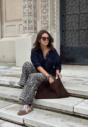 Camisa de denim con bolsillos frontales, pantalones acampanados de estampado animal, bolso tejido marrón y zapatillas marrones con cordones blancos, sentado en escalones de piedra.