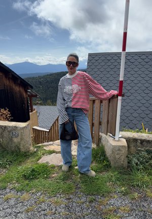 Sudadera de gran tamaño a rayas en rojo y azul marino, con texto gráfico. Combinada con jeans de color azul claro y una bolsa negra de tejido. Cabello corto; gafas de sol grandes.