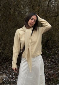 Jeune femme aux cheveux raides et foncés, vêtue d'un pull beige et d'une écharpe, portant une jupe blanche, debout en plein air avec une main sur la tête dans une zone boisée.