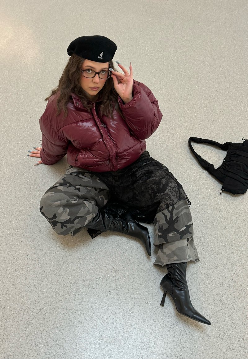 Jeune femme assise par terre portant une doudoune bordeaux, un pantalon camouflage, une jupe en dentelle noire, des bottes à talons noires, un béret noir et des lunettes, avec un sac noir à proximité.