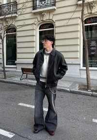 Veste en cuir noir avec un col, pull en tricot blanc, jeans foncés évasés et chaussures bordeaux. Détail de chaîne en argent sur les jeans. Cadre urbain.