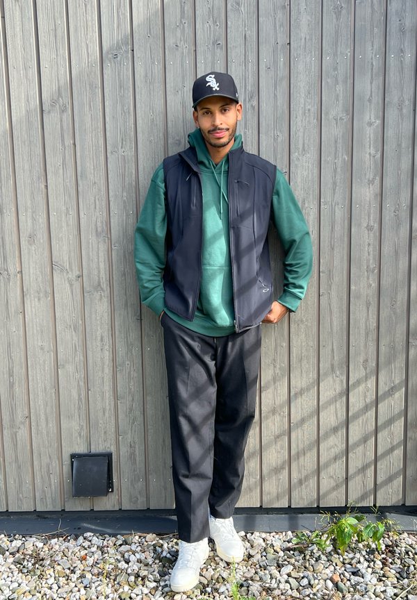 Uomo che indossa un gilet senza maniche nero sopra un hoodie verde, pantaloni neri e sneakers bianche, in piedi accanto a un muro di legno e ghiaia.