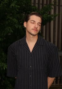 Tmavá krátkorpová košeľa s bielymi vertikálnymi pruhmi, s pevným golierom a voľným strihom, zasadená do pozadia so zeleným listnatím.