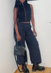 Schwarzes gestricktes, verkürztes Tanktop mit Reißverschluss, kombiniert mit lockeren schwarzen Hosen. Accessoirisiert mit einer kleinen schwarzen Handtasche und robusten schwarzen Stiefeln.