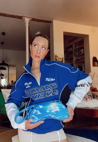 Blå lynlåst taske med sky-mønster, lavet af skinnende materiale, holdt foran en person iført en blå og hvid racertema jakke.
