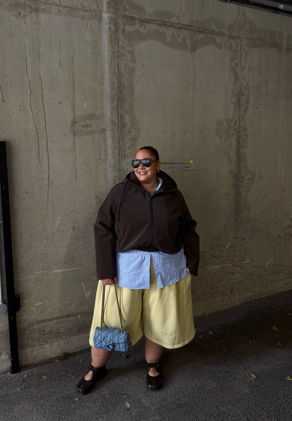 Personne portant des lunettes de soleil, une veste sombre, une chemise bleue, un short jaune et des chaussures noires, tenant un sac à main bleu, debout contre un mur en béton gris.