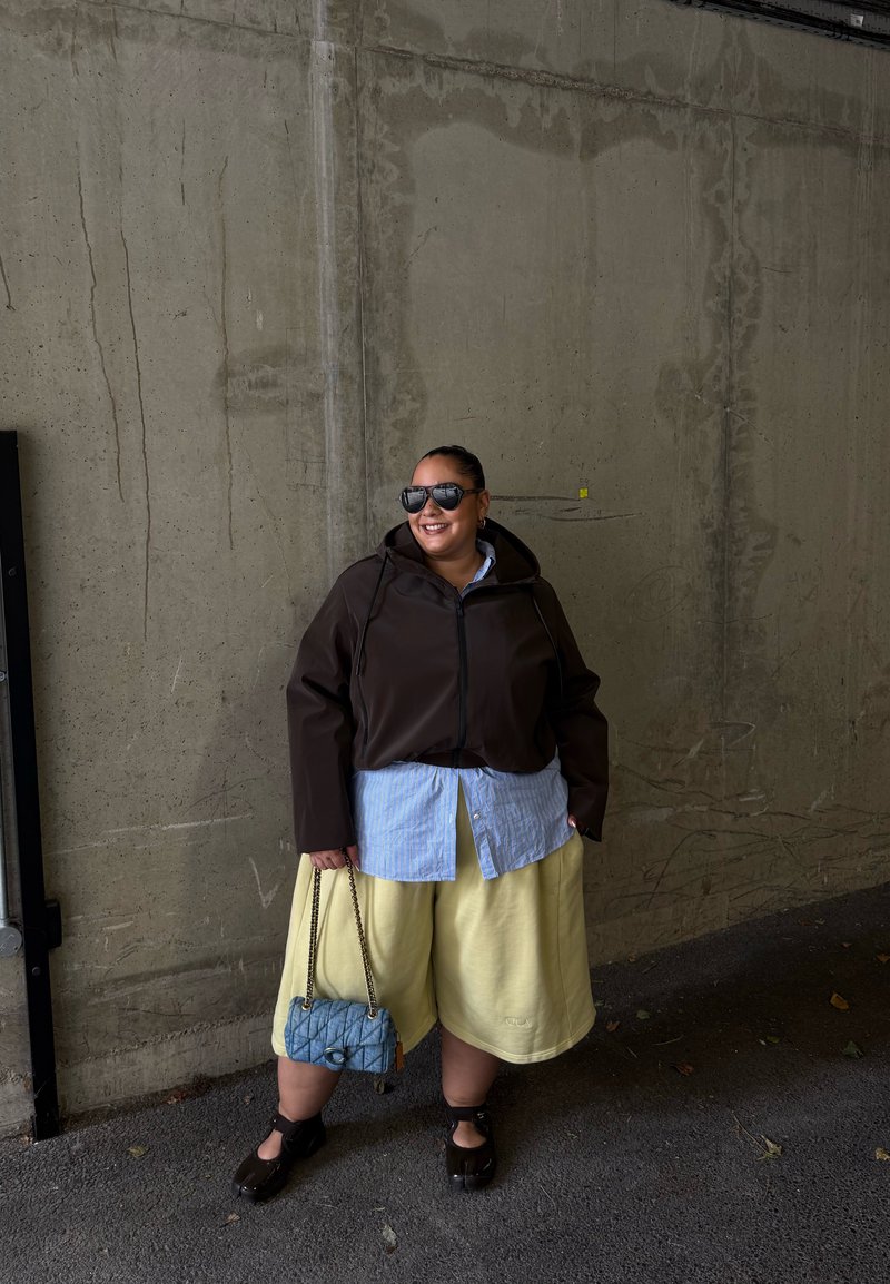 Personne portant des lunettes de soleil, une veste sombre, une chemise bleue, un short jaune et des chaussures noires, tenant un sac à main bleu, debout contre un mur en béton gris.